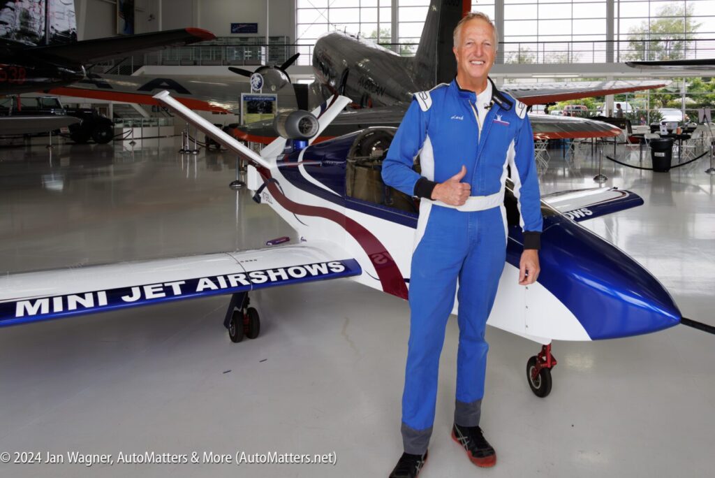 A man in a blue flight suit stands next to a small aircraft labeled "MINI JET AIRSHOWS" inside an aviation museum, giving a thumbs-up.