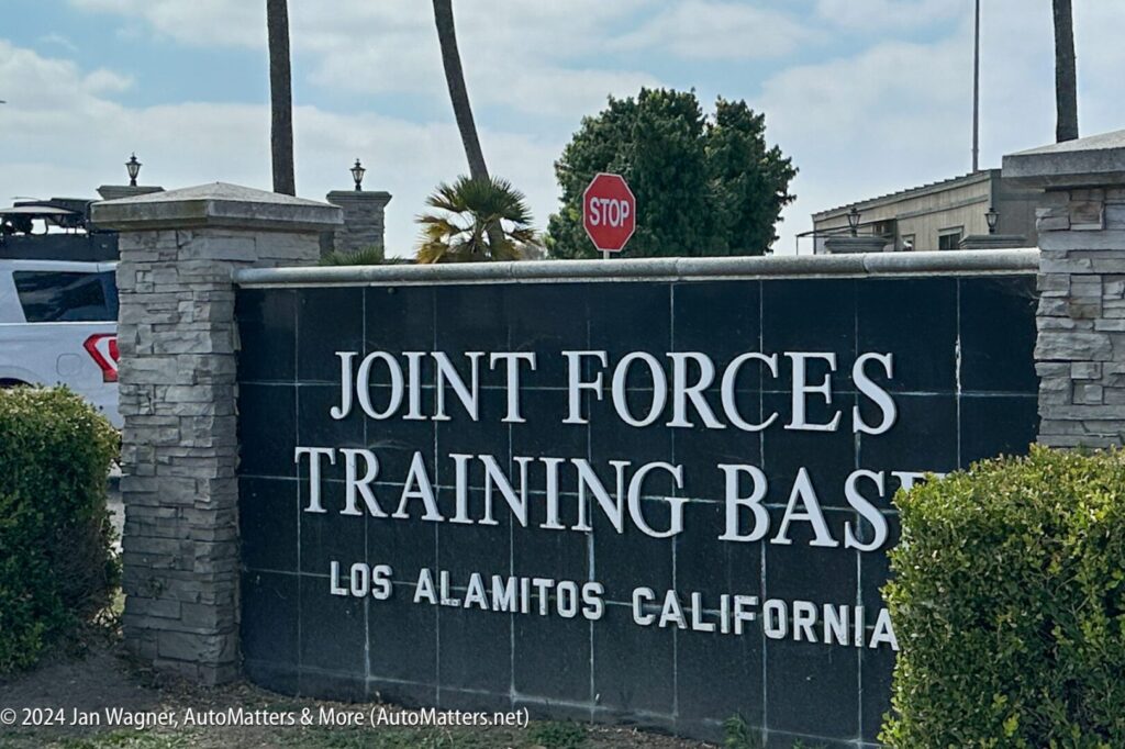 A large sign at the entrance reads "Joint Forces Training Base, Los Alamitos California," with a stone wall, plants, and a stop sign in the background.