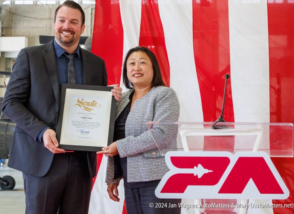A man and a woman stand in front of a red and white backdrop while the man holds a framed certificate of recognition.