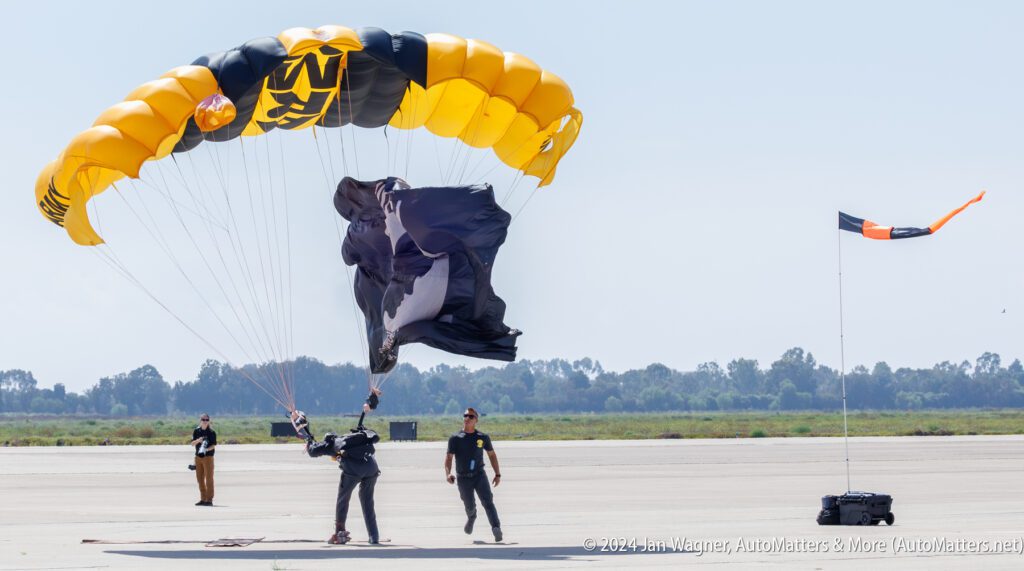 A skydiver lands on a runway with a yellow and black parachute, assisted by two people, while a wind indicator flag is visible nearby.