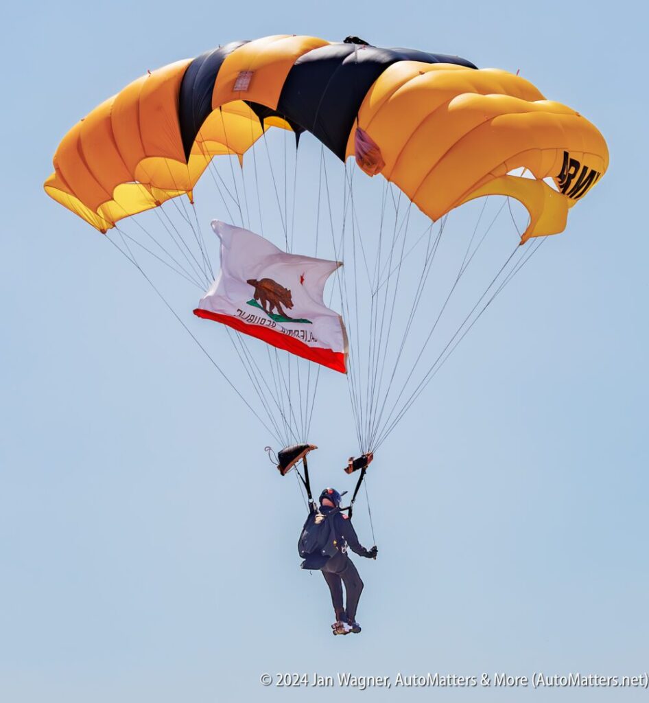 A skydiver descends with a yellow parachute, holding a California state flag against a clear blue sky.
