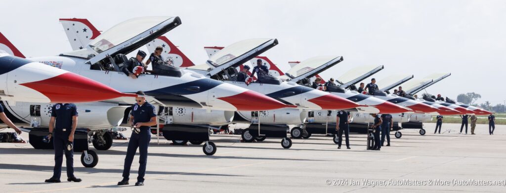 A row of fighter jets with open cockpits sits on the tarmac as pilots and crew stand nearby, preparing for flight.