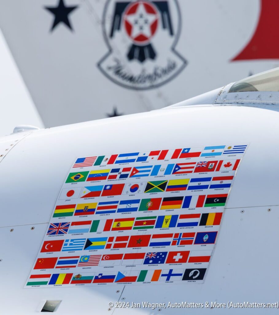 A grid of international flags is displayed on the side of a white aircraft, with part of another aircraft and insignia visible in the background.