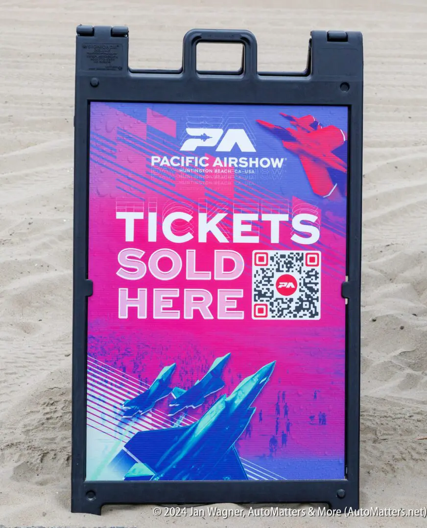A sign on sand displays "Pacific Airshow" with jet images and text reading "Tickets Sold Here" above a QR code.
