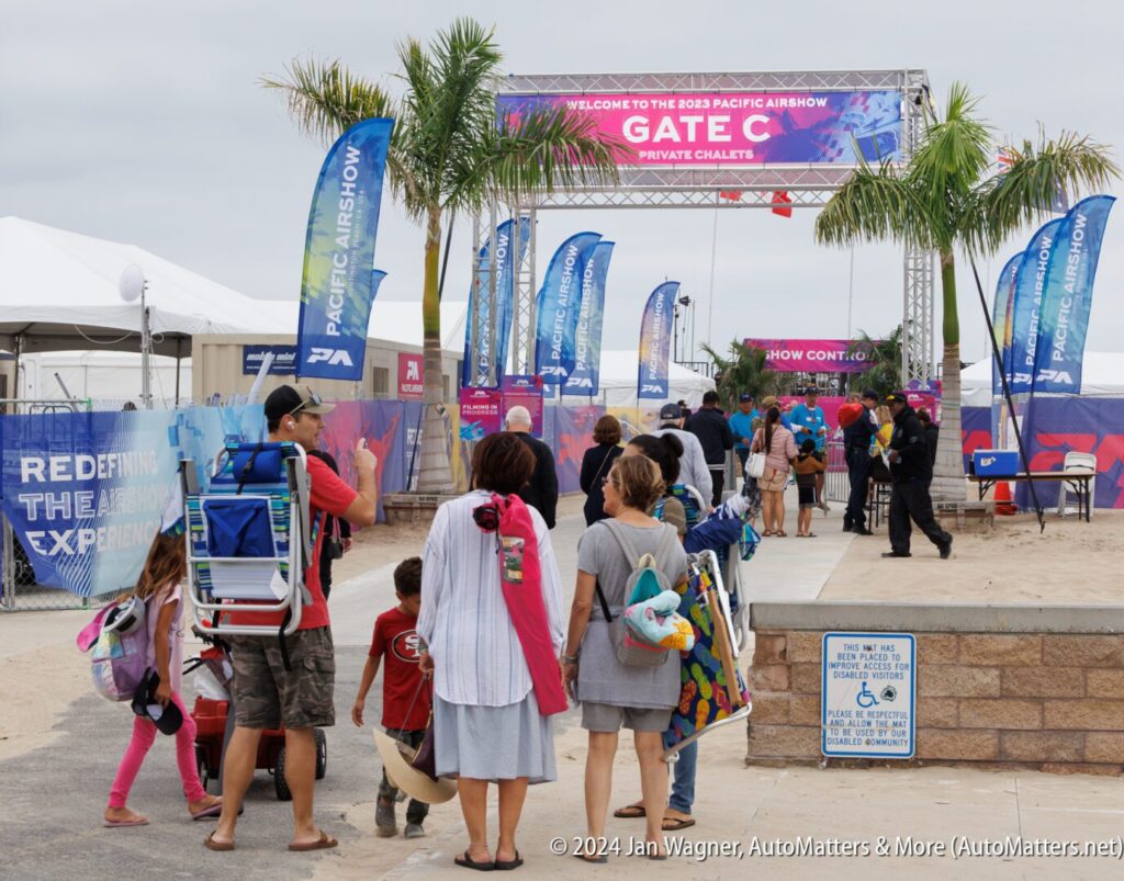 People wait in line at the entrance to the 2023 Pacific Airshow Gate C, carrying chairs and bags, with event banners and palm trees visible.