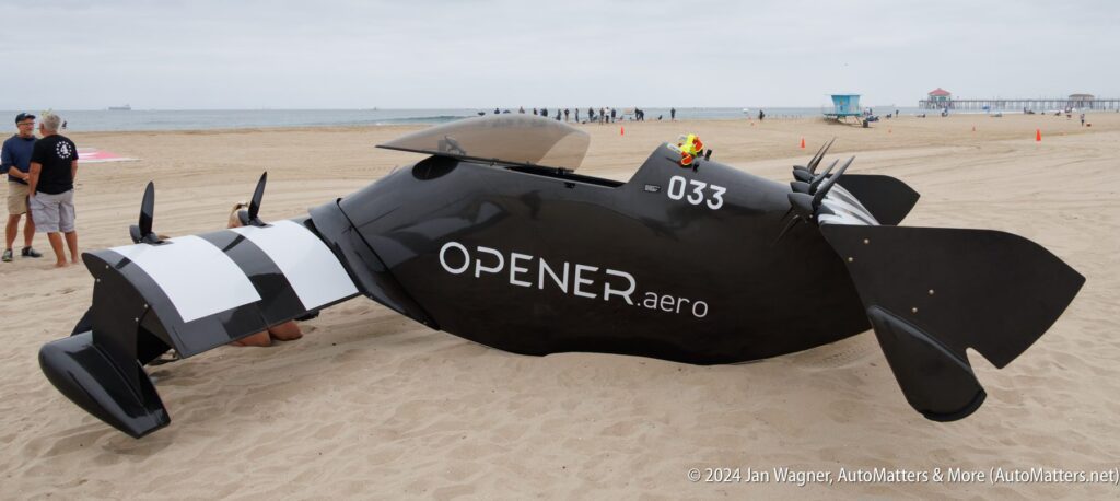 A black OPENER.aero flying vehicle with checkered accents is displayed on a sandy beach, with people and the ocean in the background.