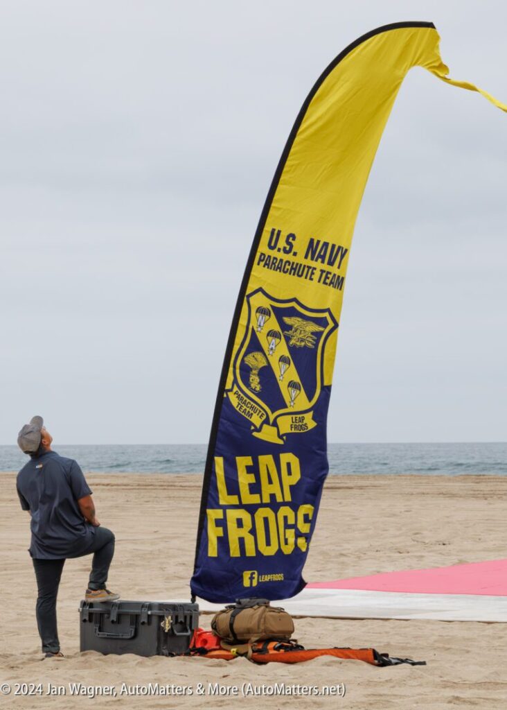 A person stands on a sandy beach near a flag reading "U.S. Navy Parachute Team Leap Frogs." Equipment cases and a large mat are also visible.