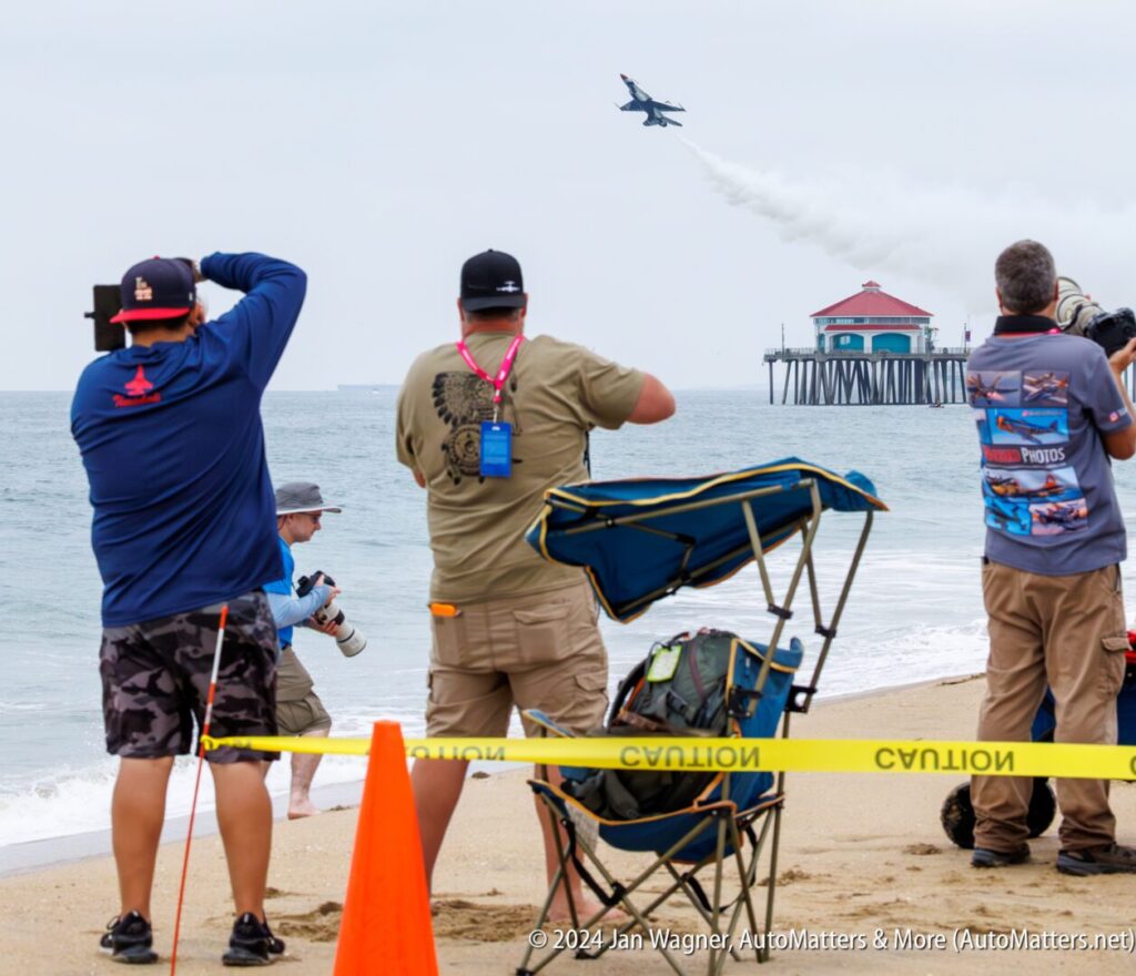 Photographers on a beach capture an airshow as a jet flies overhead, leaving a trail of smoke; caution tape marks off the area.