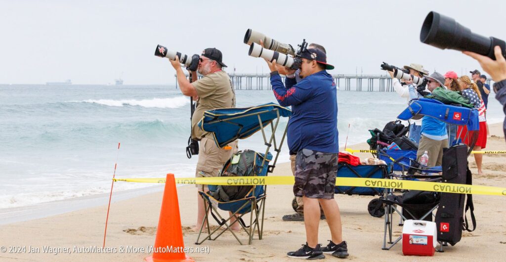 Several photographers with large cameras stand and sit behind caution tape on a sandy beach, aiming their lenses toward the ocean. A pier and oil rigs are visible in the background.