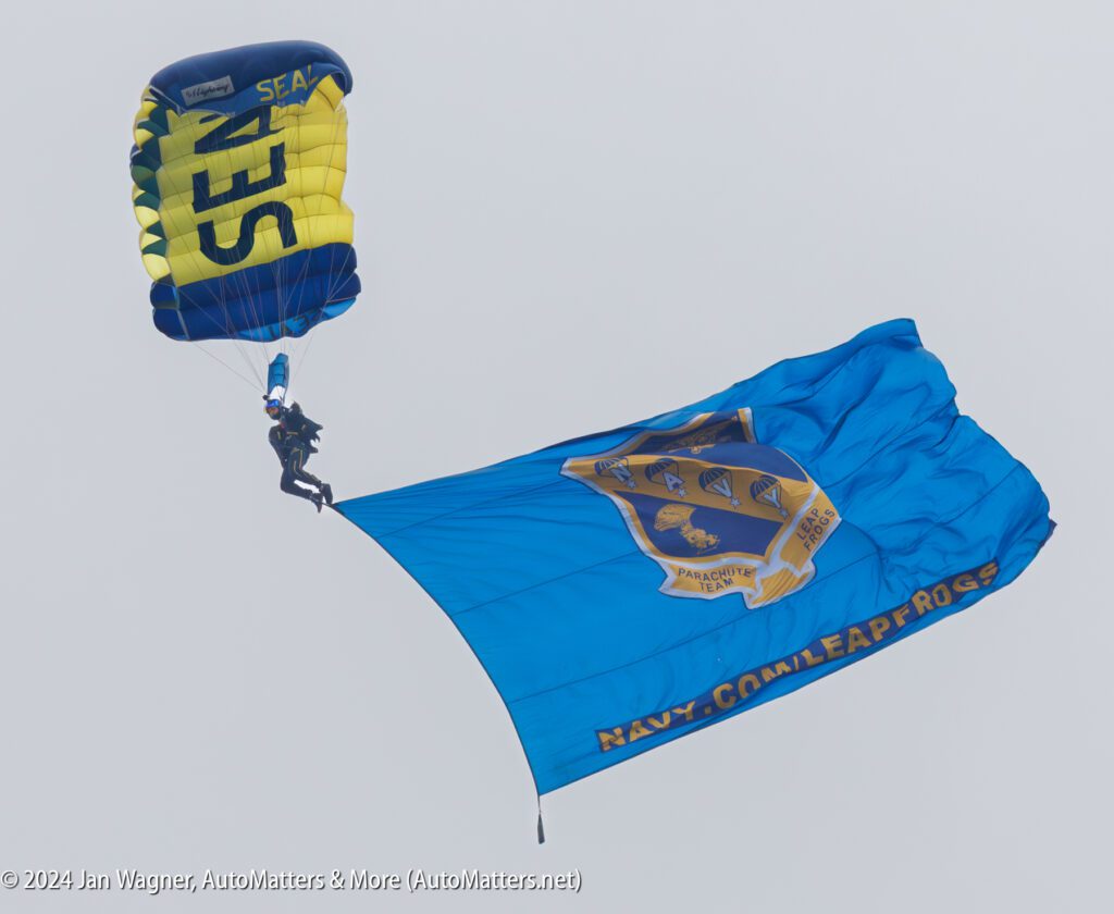 A skydiver descends with a blue and yellow parachute while displaying a large Navy-themed flag during a jump.