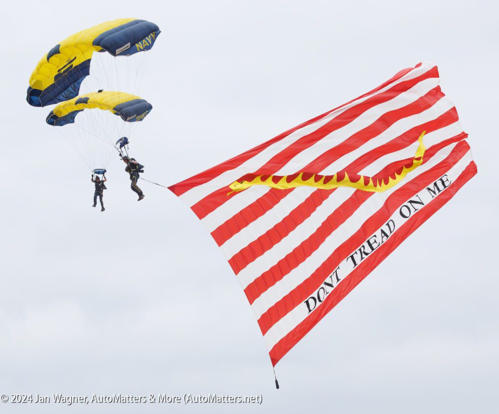 Two Navy parachutists descend with yellow-and-blue parachutes, displaying a large red-and-white striped "Don't Tread On Me" flag with a yellow snake.