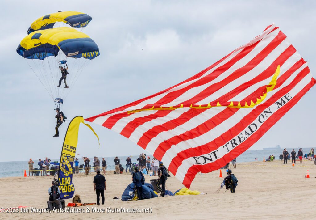 Two Navy parachutists land on a beach, one trailing a large red and white "Don't Tread On Me" flag, as a crowd watches in the background.