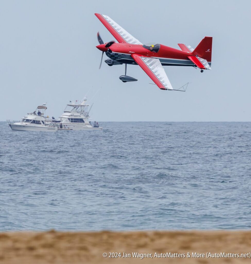 A red and white stunt plane flies low over the ocean near two boats, with sandy beach in the foreground.