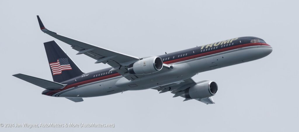 A private jet with "TRUMP" branding on the fuselage and an American flag on the tail is flying against a clear sky.