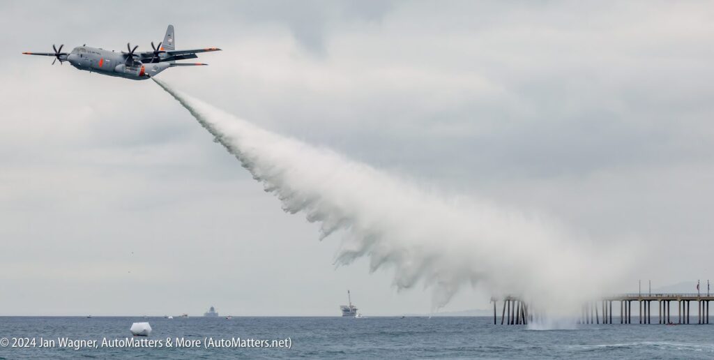 A large aircraft releases a stream of water over the ocean near a pier, creating a wide spray under a cloudy sky.