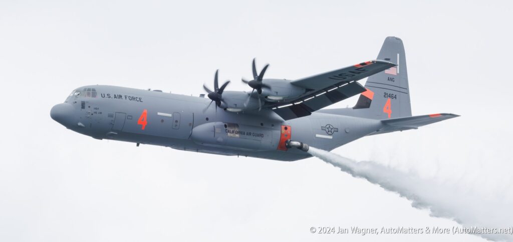 A U.S. Air Force C-130 aircraft releases a stream of water or fire retardant from its underbelly against a cloudy sky.
