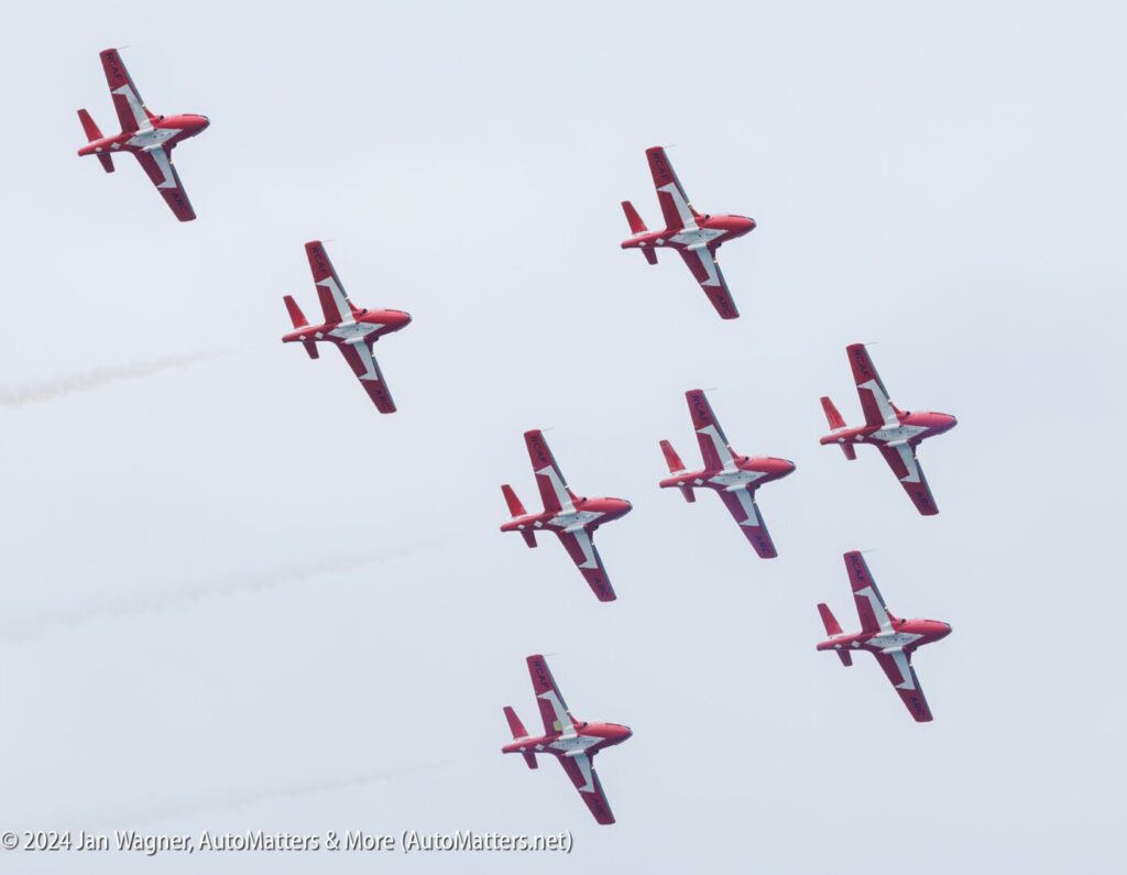 Eight red and white aircraft fly in formation against a cloudy sky, leaving faint trails of smoke behind them.