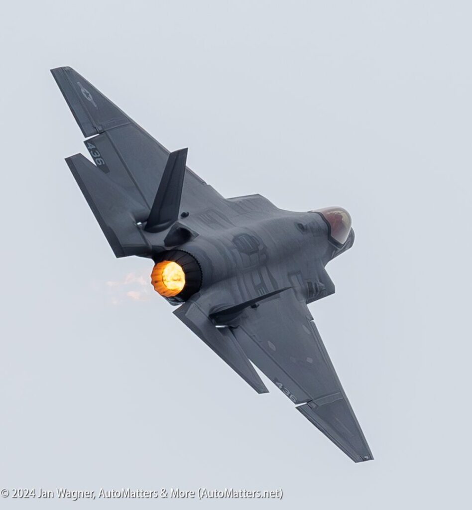 A military fighter jet with afterburner ignited is banking left in flight against a gray sky.