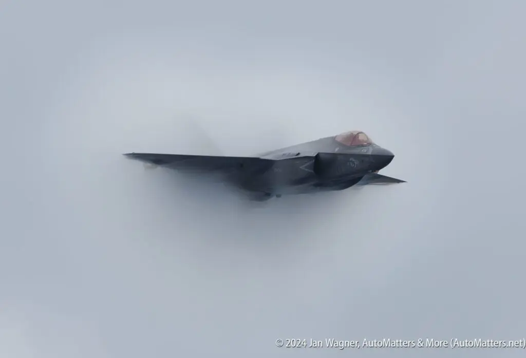 A fighter jet flies at high speed through a cloudy sky, partially obscured by vapor.