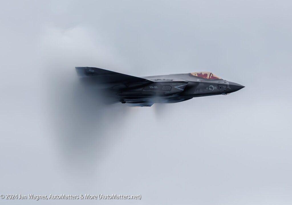 A fighter jet flies at high speed through the sky, creating a visible vapor cone around its fuselage.