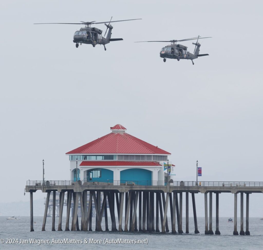 Two military helicopters fly over a pier with a building that has a red roof, above the ocean on a cloudy day.