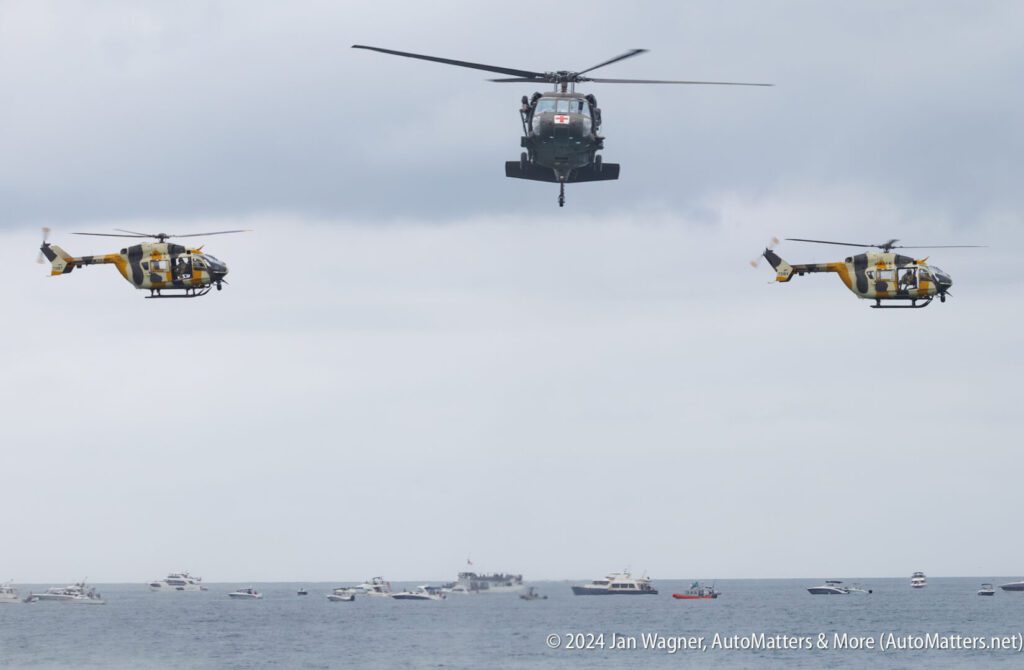 Three helicopters fly over the ocean with several boats visible below them under a cloudy sky.