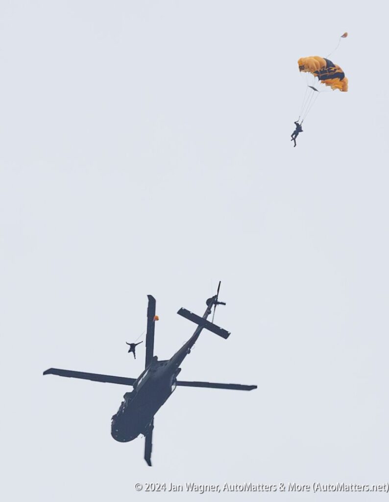 Two people parachute from a helicopter midair; one is near the open door, the other is descending with an open parachute against a pale sky.