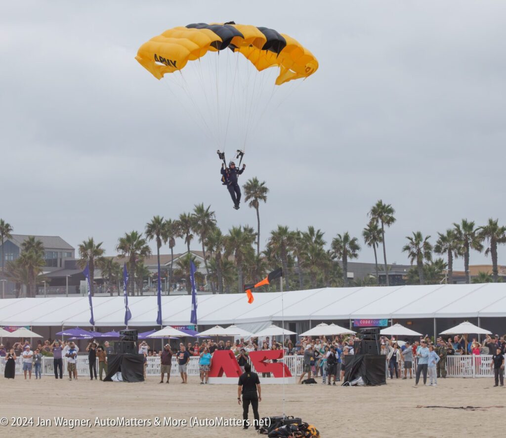 A parachutist in a yellow and black canopy prepares to land on a sandy area near a crowd at an outdoor beach event.