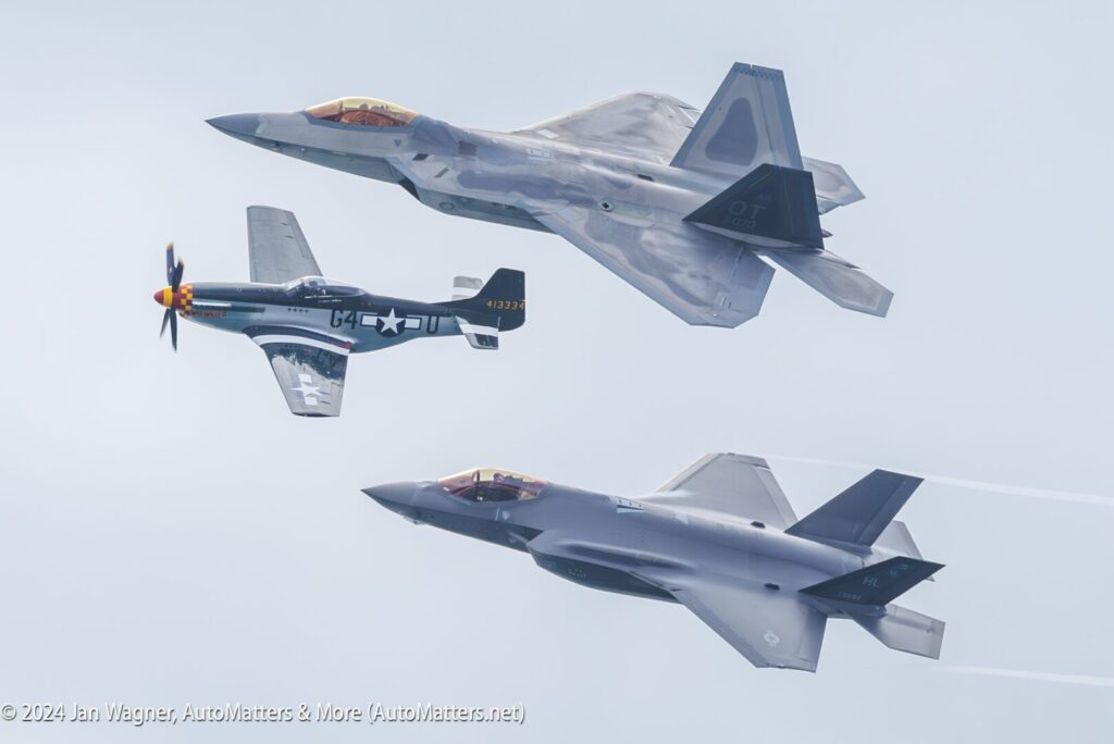 Three military aircraft fly in formation: a vintage propeller-driven fighter and two modern jet fighters, one above and one below, against a cloudy sky.