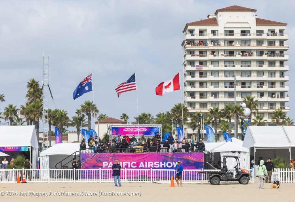 A stage setup for the Pacific Airshow on a beach with people, flags of Australia, the USA, and Canada, and a large hotel building in the background.