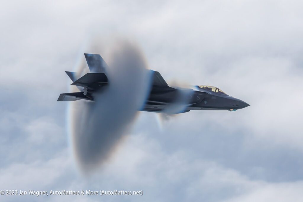 A jet fighter flies at high speed through the sky, creating a visible vapor cone around it as it approaches the speed of sound.