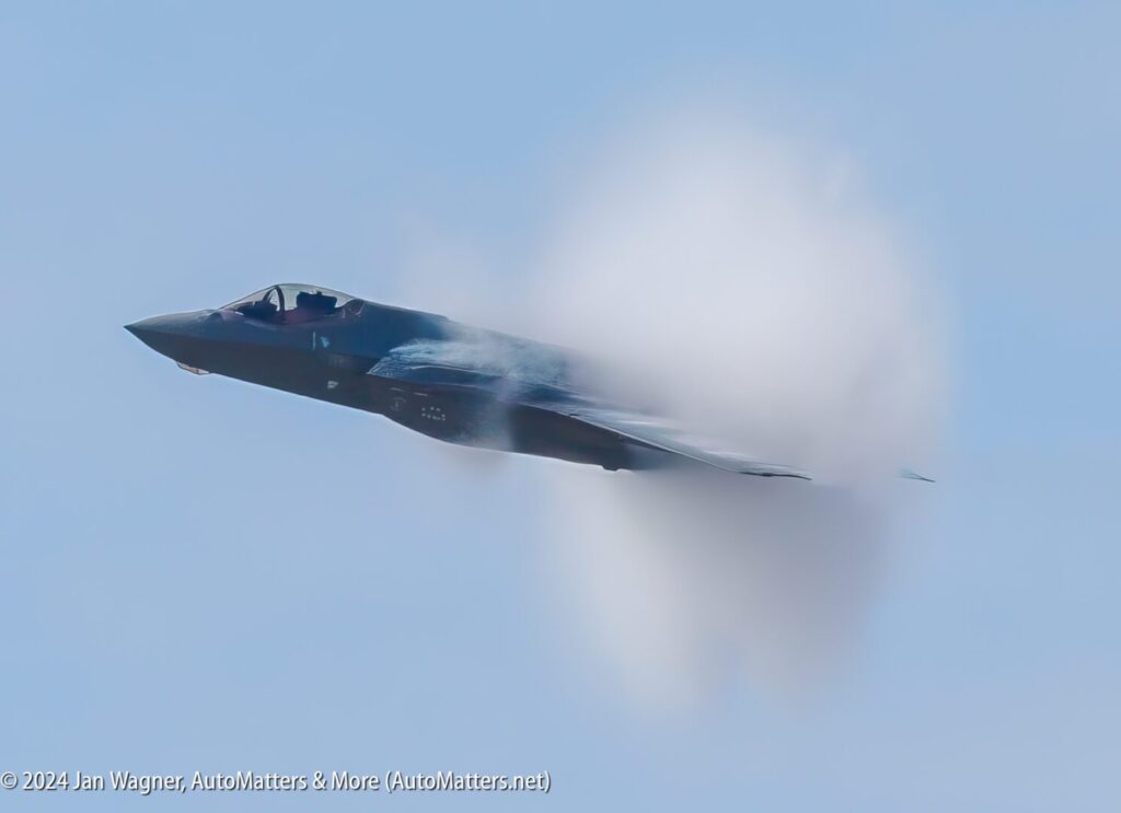 A modern fighter jet in flight, partially obscured by a vapor cone against a clear blue sky, indicating high speed.
