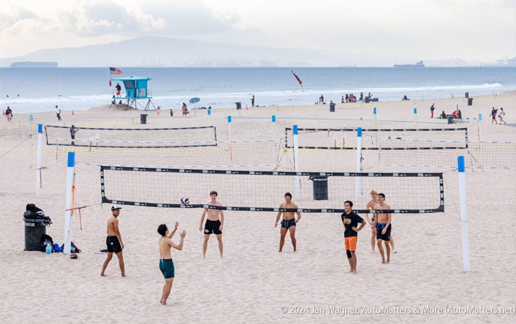 People play volleyball on a sandy beach with multiple courts set up; a lifeguard tower, beachgoers, and distant ships are visible in the background.