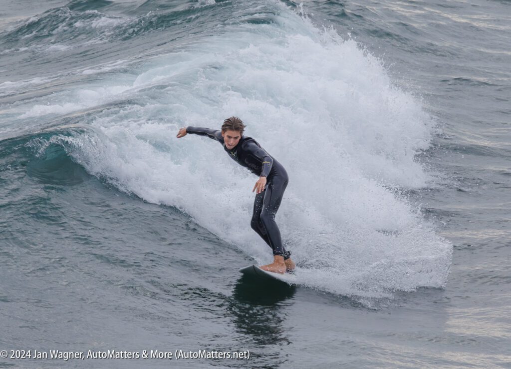 A surfer in a black wetsuit rides a wave on the ocean, maintaining balance with arms outstretched.