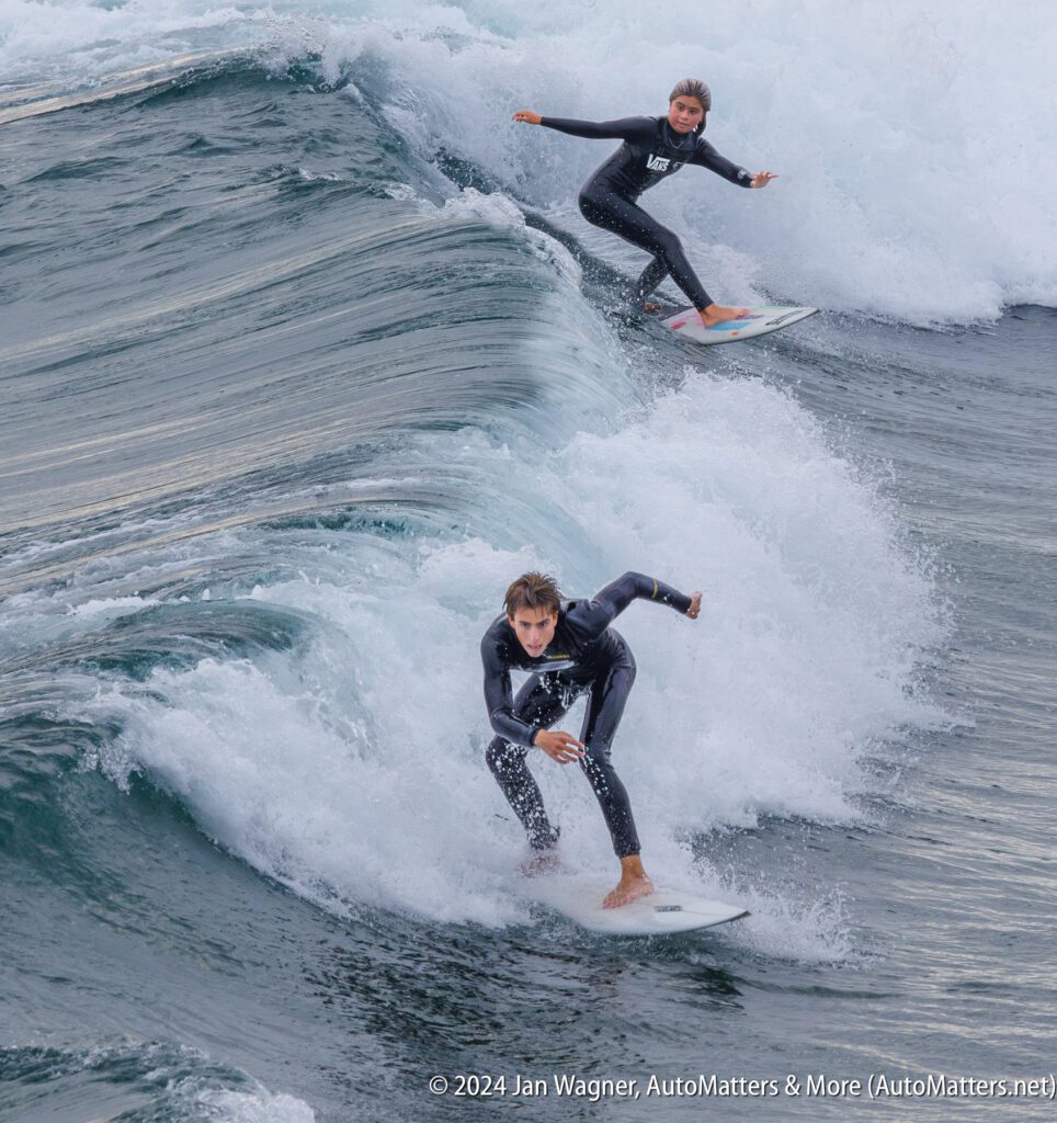 Two surfers in wetsuits ride a wave close together, with one surfer ahead and another slightly behind on the same wave.