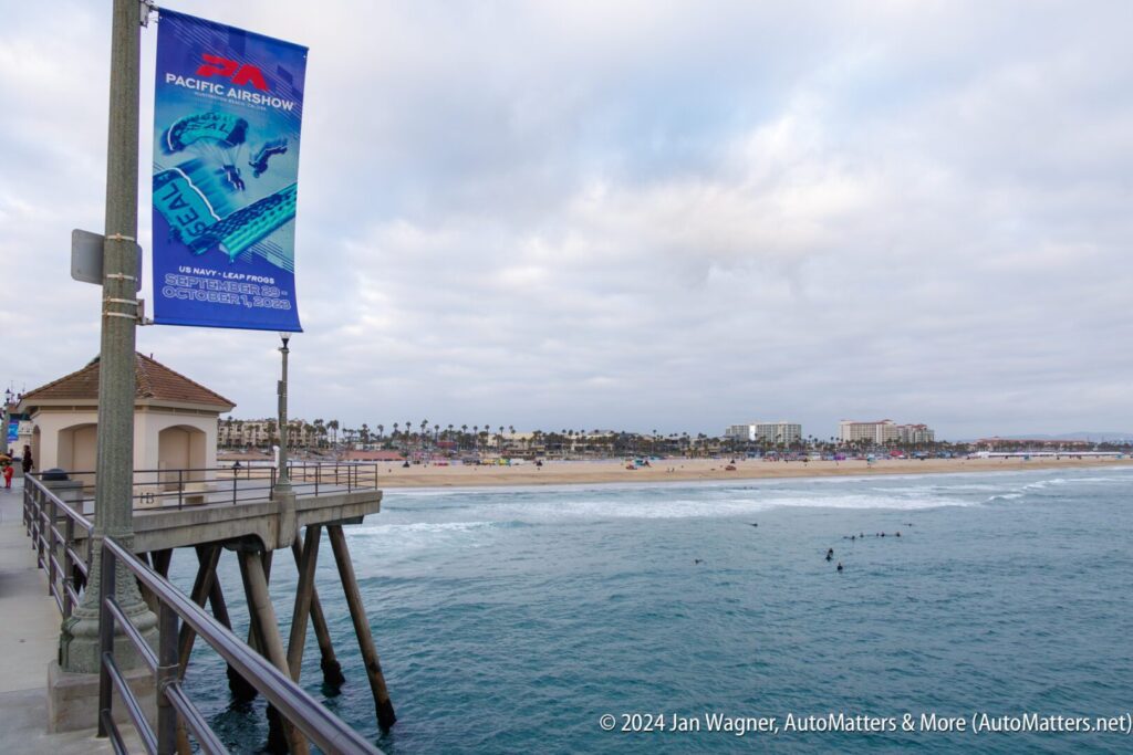 View from a pier overlooking the ocean, with a Pacific Airshow banner on a pole, a sandy beach, surfers in the water, and buildings in the background under a cloudy sky.
