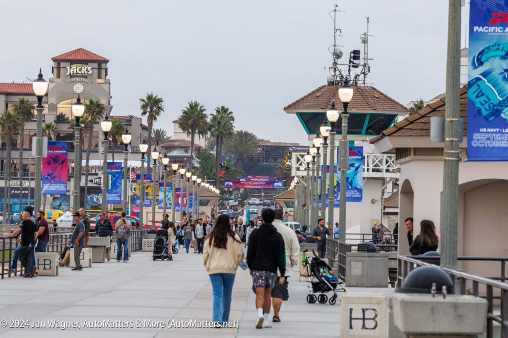 People walk along a wide pier lined with lampposts, banners, and palm trees on a cloudy day; buildings and parked cars are visible in the background.