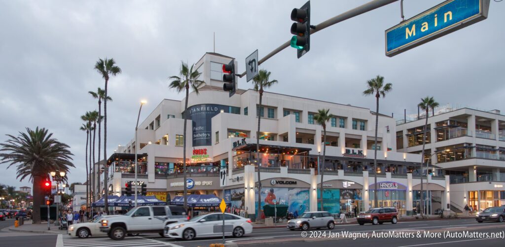 A busy street scene at dusk shows a multi-story beachside shopping and dining complex, palm trees, cars, and people near the Main Street intersection.