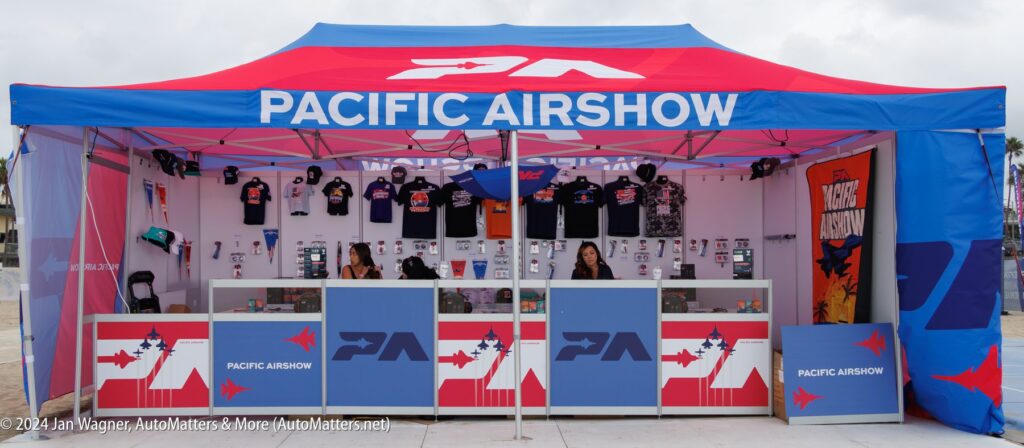 A Pacific Airshow merchandise booth with branded shirts, hats, and souvenirs on display, staffed by three people behind the counter under a blue canopy.