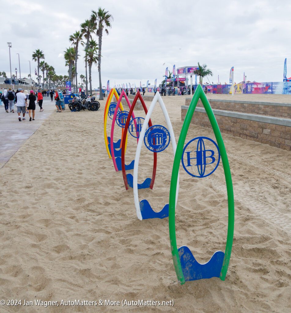Four colorful vertical surfboard sculptures with "HB" logos stand on a sandy beachside path with people, palm trees, and event tents in the background.