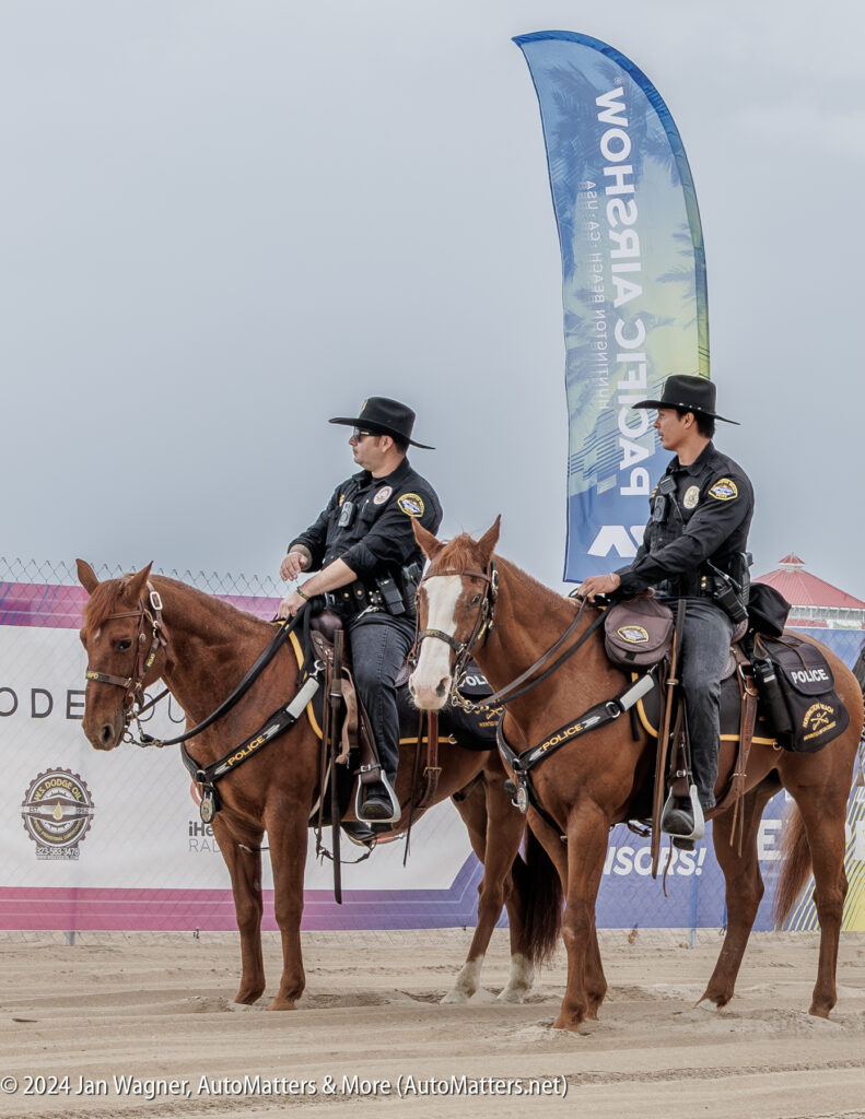 Two mounted police officers in uniform sit on brown horses at an outdoor event, with a banner and fence in the background.