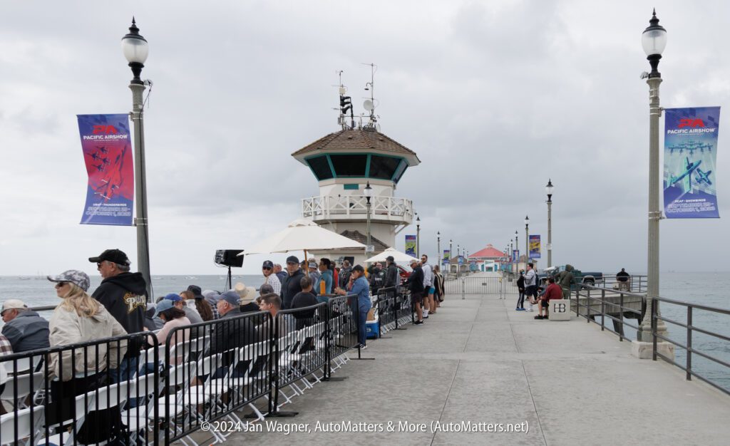 People sit and stand along a fenced area on a pier near a lifeguard tower under cloudy skies, with ocean on both sides and event banners on lamp posts.
