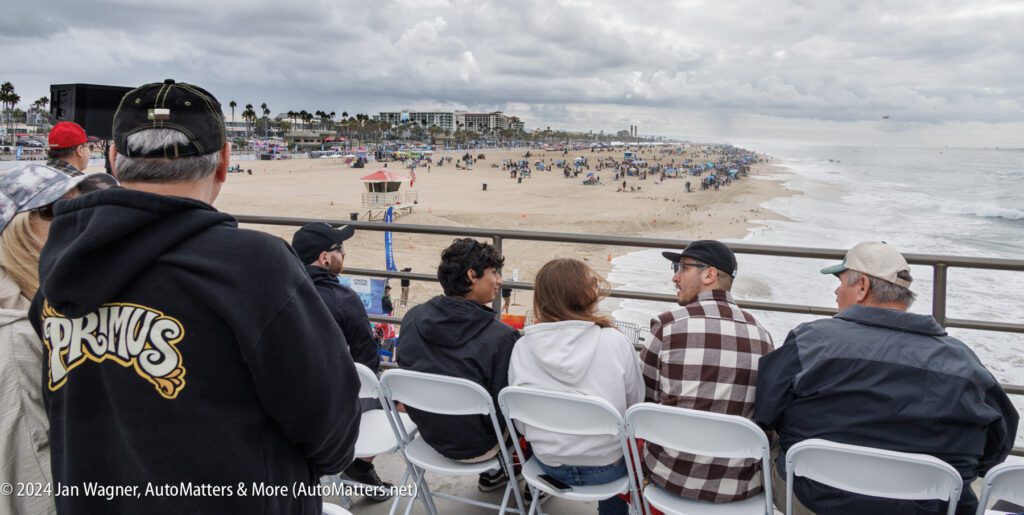 People seated on white chairs on a pier overlook a crowded beach with overcast skies and ocean waves in the background.