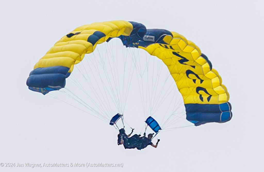 Two skydivers perform a tandem jump, descending with a yellow and blue parachute against a gray sky.