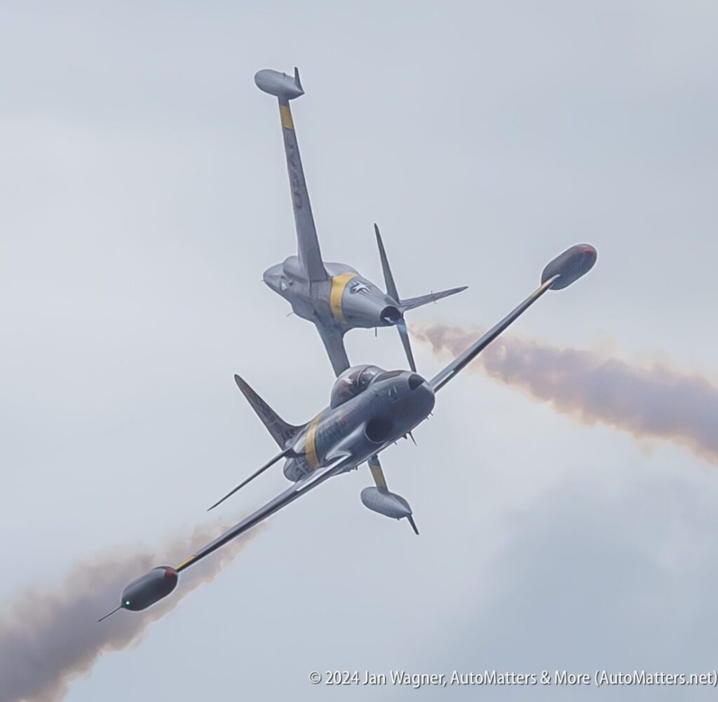 Two jet aircraft perform a close cross maneuver in the sky, trailing smoke against a cloudy background.