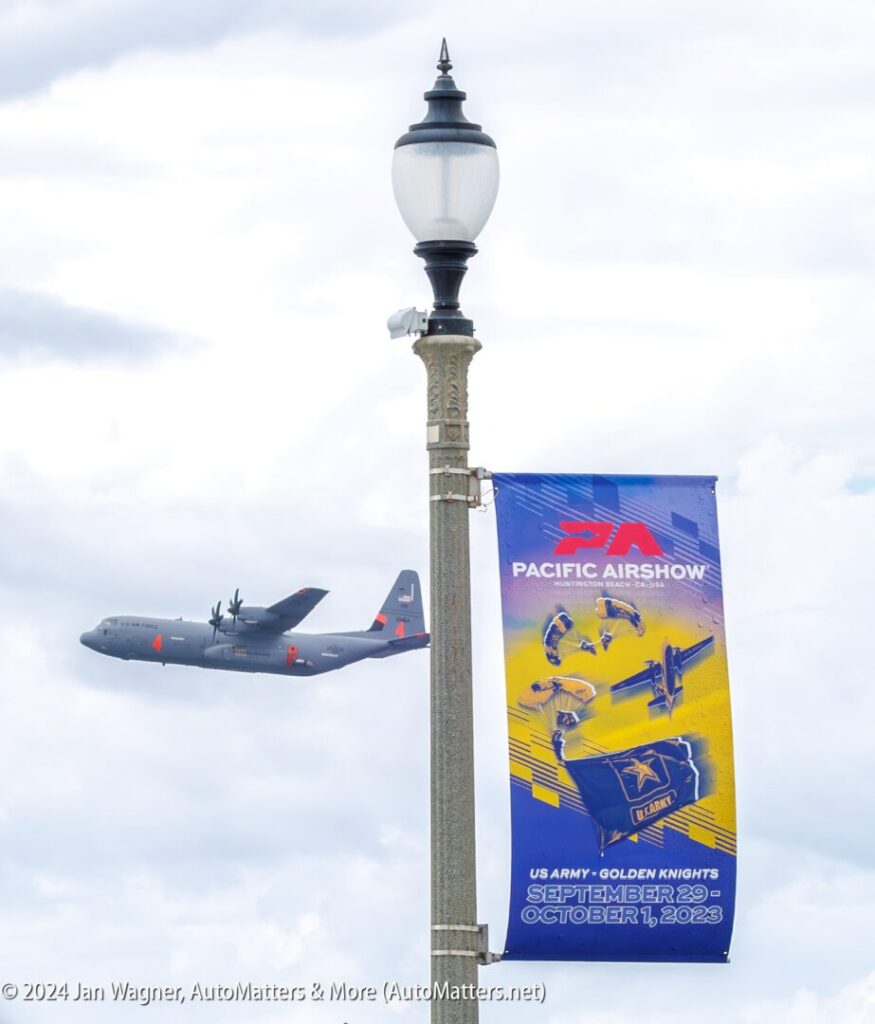 A military aircraft flies in the sky near a streetlamp with a banner advertising the Pacific Airshow, scheduled for September 29 to October 1, 2023.