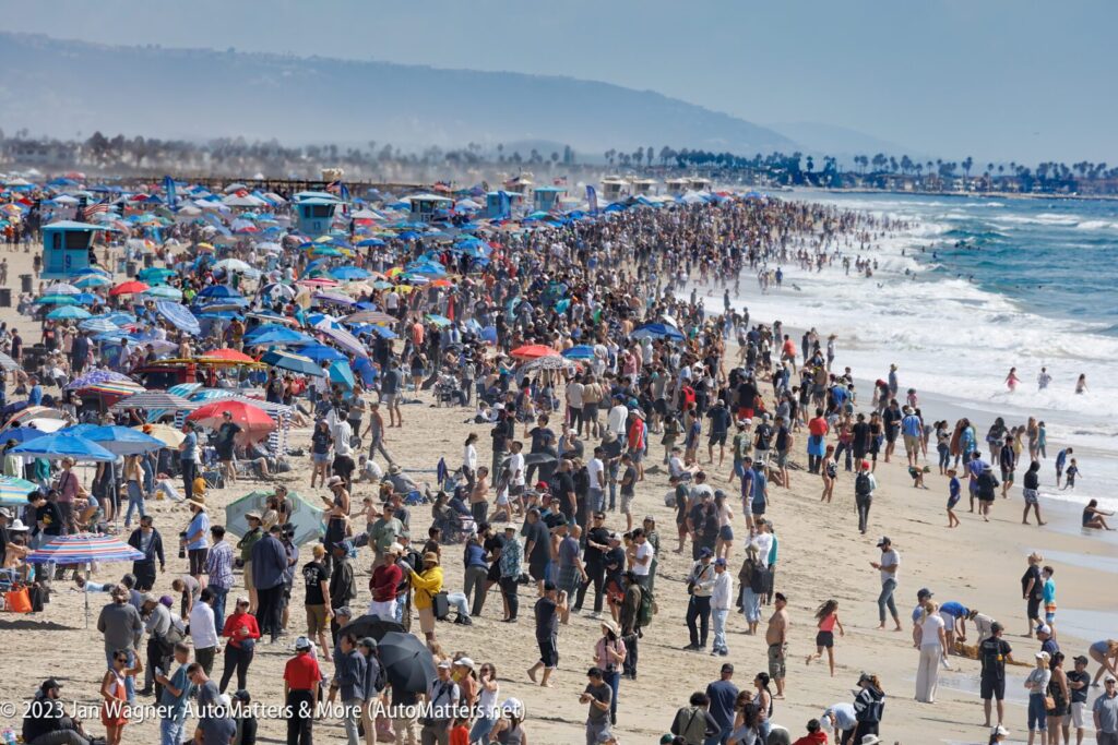 A crowded beach scene with hundreds of people, colorful umbrellas, and lifeguard towers along the shoreline on a bright, sunny day.