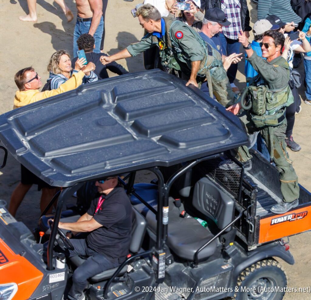 Two people in flight suits greet a crowd from the back of a utility vehicle on a sandy area, while a driver sits in the front seat.