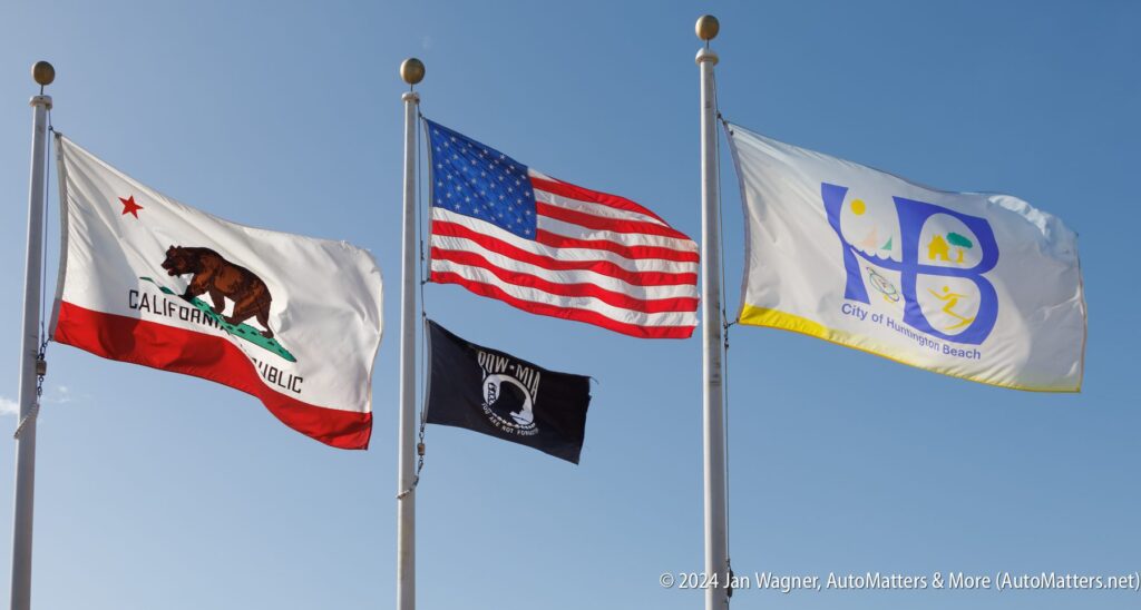 Four flags flying against a clear blue sky: California state flag, U.S. flag, POW/MIA flag, and City of Huntington Beach flag.