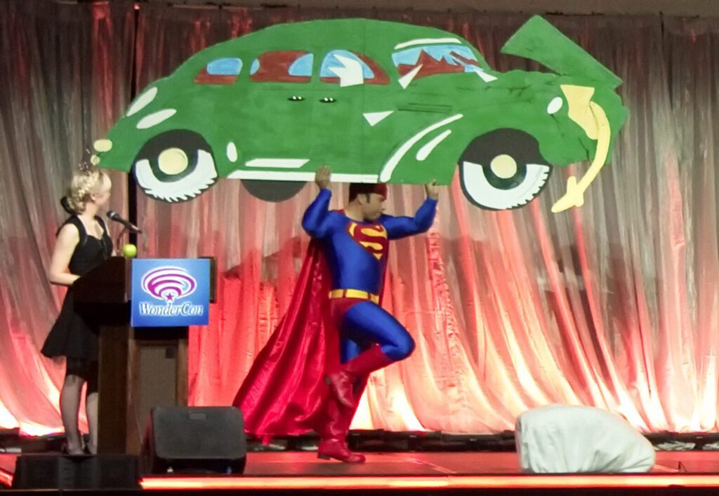 A man in a superman costume is holding a car on stage.